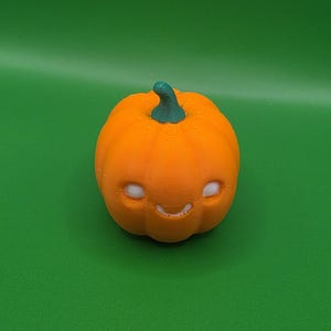 Könnte beinhalten: Eine kleine, orangefarbene, 3D-gedruckte Kürbisfigur mit einem grünen Stiel. Der Kürbis hat eine glatte, abgerundete Form mit vertikalen Rillen und ein einfaches, geschnitztes Gesicht, das aus zwei weißen runden Augen und einem kleinen, geschwungenen Mund besteht.