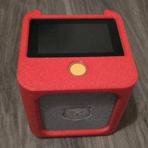 Könnte beinhalten: Ein quadratisches, rotes elektronisches Gerät mit einem schwarzen Bildschirm, einem gelben runden Knopf und einem grauen Lautsprechergitter, das ein kleines zwinkerndes Katzensymbol zeigt. Das Gehäuse ist texturiert und hat abgerundete Ecken.