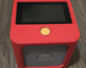 Könnte beinhalten: Ein quadratisches elektronisches Gerät mit einem leuchtend roten Silikongehäuse, das einen schwarzen rechteckigen Bildschirm auf der Oberseite, einen einzelnen gelben runden Knopf und ein graues Lautsprechergitter mit einem zwinkernden Katzensymbol besitzt.