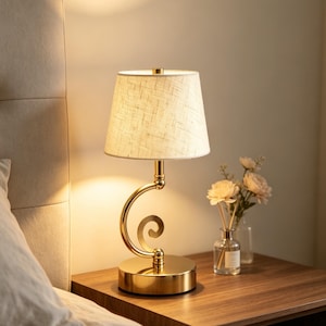 May include: A table lamp with a polished gold-tone metal base featuring a curved, spiral-shaped neck and a tapered, off-white fabric lampshade with a visible woven texture. The lamp emits a warm, soft glow, illuminating the surrounding area.