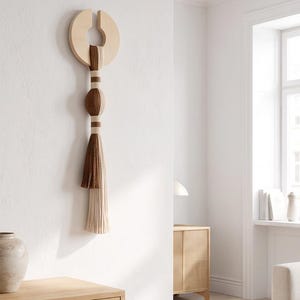 May include: A wall hanging featuring a light-colored wooden circular frame with a gap at the top. Suspended from the frame is a vertical arrangement of yarn in shades of cream, tan, and dark brown, featuring a central bulbous knot.