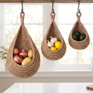 May include: Three teardrop-shaped hanging baskets made of woven tan jute rope. Each basket is suspended from a dark metal hook. The baskets contain various produce: red onions and potatoes in the largest, garlic and lemons in the middle, and green avocados in the smallest.