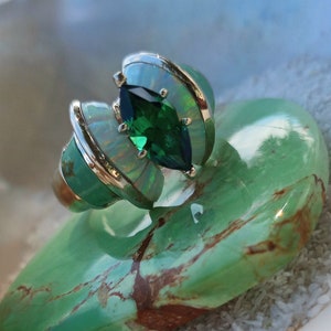 May include: A silver ring with a green gemstone and opal accents. The ring is set on a green stone background.