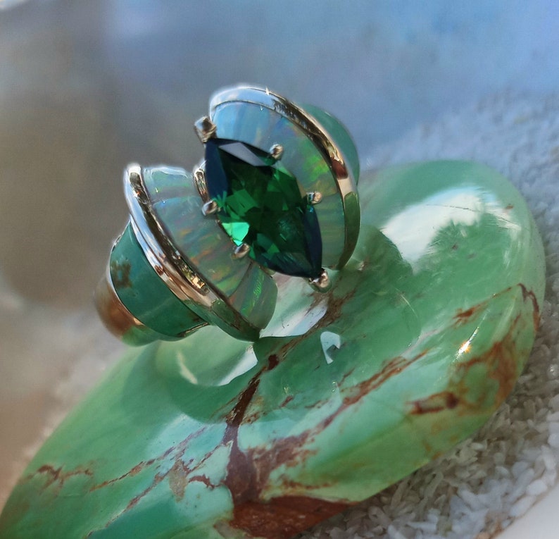 May include: A silver ring with a green gemstone and opal accents. The ring is set on a green stone background.