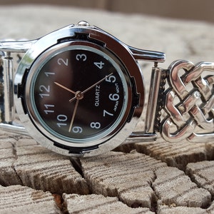 May include: A silver watch with a black face and a Celtic knot design band. The watch face has black Roman numeral hour markers and a quartz movement. The text "JAPAN MVT" is printed on the watch face.