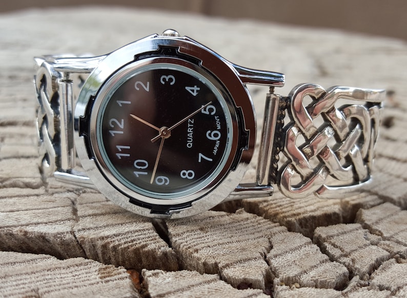 May include: A silver watch with a black face and a Celtic knot design band. The watch face has black Roman numeral hour markers and a quartz movement. The text "JAPAN MVT" is printed on the watch face.