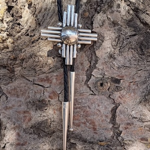 Puede incluir: Un bolo tie de plata con un diseño geométrico en forma de estrella. El bolo tie está en un cordón de cuero negro y tiene una punta de plata.