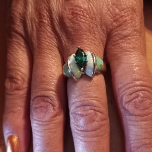 May include: A silver ring with a green gemstone and white opal accents. The ring is on a person's finger.