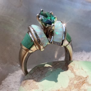 May include: A silver ring with a green gemstone and white opal accents. The ring is set on a light green surface.