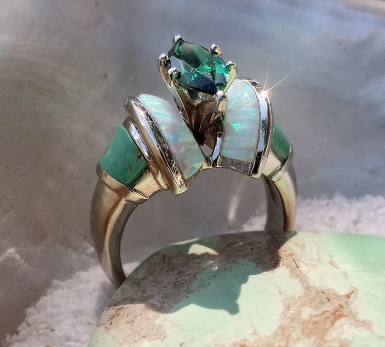 May include: A silver ring with a green gemstone and white opal accents. The ring is set on a light green surface.