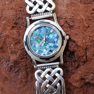 May include: A silver watch with a Celtic knot design band and a face made of iridescent opal mosaic tiles. The watch face has black markings and says "QUARTZ" at the bottom.