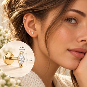 Può includere: Un orecchino a lobo in oro con retro piatto e una pietra preziosa trasparente taglio marquise. L'orecchino misura 1,5 mm di larghezza e 3 mm di altezza, come indicato dal testo e dalle linee. È mostrato in un riquadro circolare e indossato.