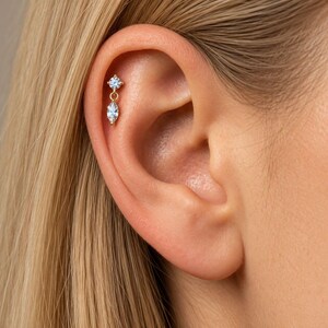 May include: A gold cartilage earring featuring a small round clear stone stud connected by a thin gold chain to a marquise-cut clear stone drop. The jewelry is positioned in the upper ear cartilage, showcasing a delicate metal design.