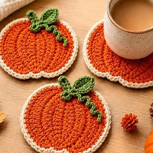 May include: Two round, orange, crocheted pumpkin-shaped coasters with a cream-colored border. Each coaster features a green crocheted leaf and a curly vine detail on top. One coaster is placed under a ceramic mug filled with a light brown beverage.