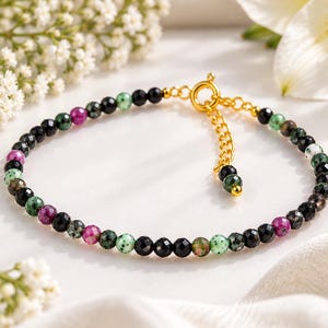 Pode incluir: Uma pulseira de contas com pequenas pedras preciosas redondas e facetadas em preto, magenta e verde salpicado. A pulseira inclui um fecho de mola em metal dourado e uma corrente de extensão ajustável com uma conta na extremidade.