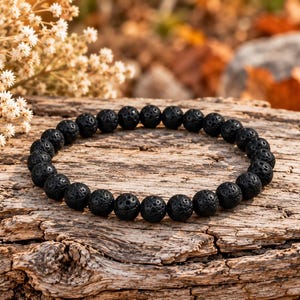 Könnte beinhalten: Ein kreisförmiges Armband, bestehend aus zahlreichen kleinen, kugelförmigen schwarzen Lavaperlen mit einer porösen, texturierten Oberfläche. Die Perlen sind zu einer durchgehenden Schlaufe aufgereiht und zeigen die natürliche, unebene Struktur.
