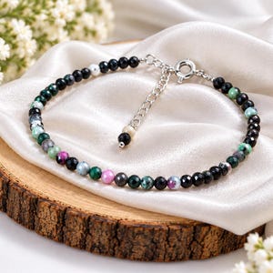 Könnte beinhalten: Ein zartes Armband mit kleinen, facettierten, mehrfarbigen Edelsteinperlen in Schwarz, Grün, Rosa und Weiß. Das Armband verfügt über eine silberfarbene Metallverlängerungskette mit einem runden Verschluss und einer kleinen Perle am Ende.