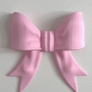 May include: A light pink bow with a matte finish and a visible 3D printed texture. The bow features two large loops and two downward-pointing tails with a central knot detail, creating a structured and symmetrical decorative object.