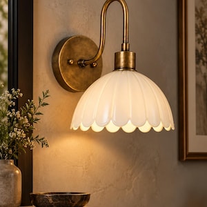 Könnte beinhalten: Eine Wandleuchte mit einem geschwungenen Metallarm in Antikmessing-Optik und einer runden Wandplatte. Die Lampe verfügt über einen gewellten, weißen Glasschirm mit blütenblattartigem Rand, der das Licht von der Messingfassung nach unten lenkt.