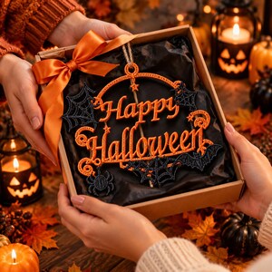 Op de afbeelding: Een decoratief hangornament in een geschenkdoos met de oranje geborduurde tekst Happy Halloween, geaccentueerd met zwarte spinnenwebben, een spin en een vleermuis, opgehangen aan een touwtje van jute en gestrikt met een oranje satijnen lint.
