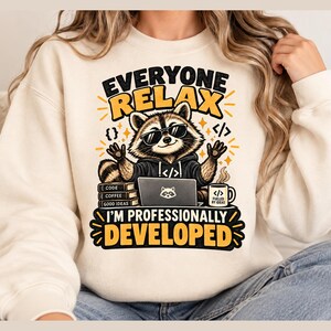 May include: A cream-colored crewneck sweatshirt featuring a graphic of a raccoon wearing sunglasses, sitting at a laptop with books and a coffee mug. The text reads: EVERYONE RELAX I'M PROFESSIONALLY DEVELOPED, CODE, COFFEE, GOOD IDEAS, and FUELED BY IDEAS.
