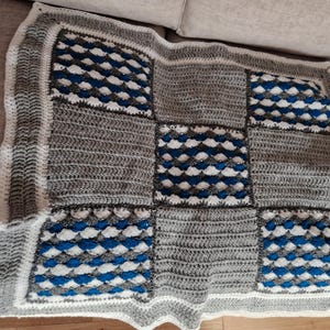 May include: A square crochet blanket featuring a patchwork design of alternating grey textured panels and blue, white, and grey shell-stitch squares. The blanket is finished with a white border and a grey ribbed edge, creating a geometric pattern.