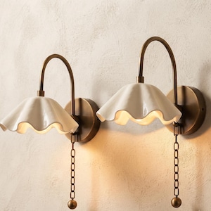 Puede incluir: Dos lámparas de pared con brazos curvos de latón antiguo y placas circulares. Cada lámpara tiene una pantalla de cerámica blanca festoneada con forma de pétalo de flor. Una cadena de metal con un peso esférico cuelga de cada accesorio.