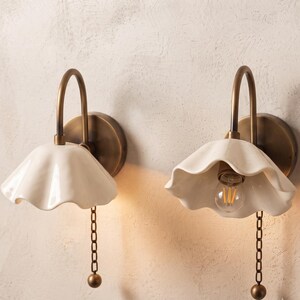 Peut inclure: Deux appliques murales dotées de bras courbes en laiton antique et de plaques arrière circulaires. Chaque lampe possède un abat-jour en céramique blanc cassé à bords ondulés et une chaînette avec un poids sphérique. Une lampe est allumée.