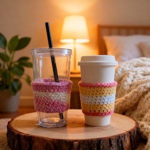 Puede incluir: Un vaso de plástico transparente con pajita negra y funda de ganchillo rosa, blanca y amarilla, junto a un vaso blanco con tapa y funda de ganchillo amarilla, blanca y rosa, ambos sobre una rodaja de madera circular.