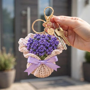 May include: A crochet keyring featuring a bouquet of purple lavender flowers wrapped in beige yarn, tied with a light purple satin ribbon. The keyring includes a gold-toned metal clasp, a ring, and a small round tag engraved with "hand made" and a heart.