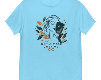 Könnte beinhalten: Ein hellblaues T-Shirt mit einer grafischen Illustration einer Person, die eine dunkle Maske hält. Das Design enthält orangefarbene und dunkelgrüne Blätter, Sterne, eine Mondsichel und ein Unendlichkeitssymbol. Der Text lautet: NOT A MASK. JUST ME.