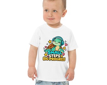 Könnte beinhalten: Ein weißes Kurzarm-T-Shirt mit einer Grafik einer grünen Schildkröte, die einen blauen Kopfhörer trägt. Das Design enthält gelbe Sterne, rosa Herzen und den Text "SMALL STEPS BIG PROGRESS" in blauer und gelber Schrift auf der Vorderseite.