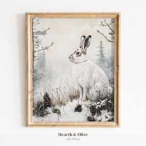 May include: A framed art print featuring a white hare sitting in a snowy landscape with pine trees and scattered pinecones. The artwork is enclosed in a light-coloured wooden frame. The text Hearth & Olive Art Prints is visible below.