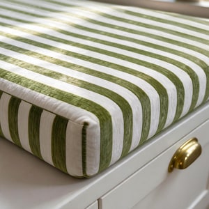 May include: A rectangular bench cushion featuring a vertical striped pattern in olive green and white. The fabric has a soft, velvet-like texture. The cushion sits atop a white wooden base equipped with a polished brass cup drawer pull.