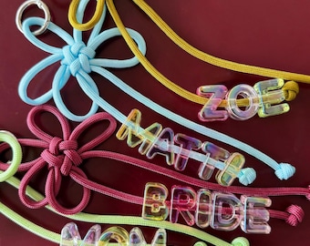 Op de afbeelding: Vier bloemvormige sleutelhangers gemaakt van koord in mosterdgeel, lichtblauw, donkerroze en limoengroen. Elke sleutelhanger heeft iriserende, doorschijnende plastic letterkralen die de namen ZOE, MATT, BRIDE en MOM spellen.
