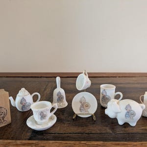 Peut inclure: Une collection d'objets miniatures en céramique blanche ornés d'illustrations Precious Moments, comprenant une théière, des tasses avec soucoupes, une cloche, une tirelire, un petit sac et un bloc en bois, décorés de dessins aux tons doux.