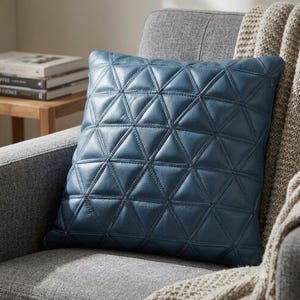 May include: A square blue leather throw pillow featuring a geometric pattern of stitched triangles across the front surface. The leather has a visible grain texture and is constructed from multiple triangular panels joined by visible seams.