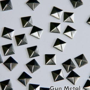 May include: A close-up of a pile of silver pyramid-shaped studs. The studs are scattered on a white surface. The text "Gun Metal/Dark Silver" is visible at the bottom of the image.