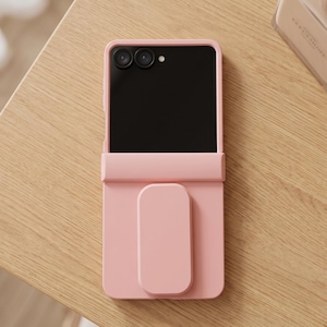 May include: A light pink smartphone case featuring a smooth matte finish and a rectangular grip attachment on the lower half. The case covers the back and sides of the device, leaving the dual camera lenses and flash exposed at the top section.