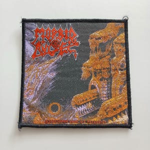 May include: A square fabric patch featuring the Morbid Angel logo in red text at the top left. The design includes a dark background with purple and orange abstract imagery. The text at the bottom reads 2000 MORBID ANGEL ULE RAZAMATAZ.