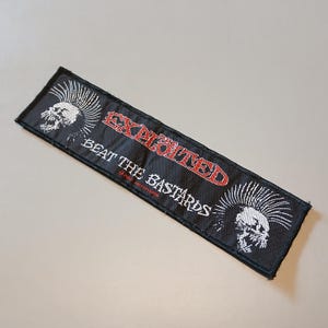 May include: A rectangular black embroidered patch featuring the text EXPLOITED in red and BEAT THE BASTARDS in white. The design includes two white skull graphics with mohawk hairstyles on the left and right sides of the fabric patch.