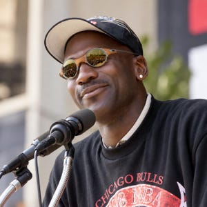 May include: A person wearing a black baseball cap with white and red accents, and round sunglasses with gold-tinted lenses. The person is wearing a black sweatshirt with the text Chicago Bulls and NBA printed in red and white on the front.