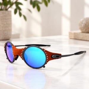 May include: Sunglasses featuring round, reflective blue lenses set within black frames. The bridge and outer edges are wrapped in a textured, rust-coloured material secured with visible, light-coloured stitching. The temples are thin, black, and straight.
