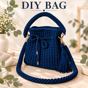 Puede incluir: Un bolso tipo cubo de ganchillo azul oscuro con un asa trenzada gruesa y una correa larga para el hombro. El bolso cuenta con un cierre de cordón con puntas de metal dorado y un accesorio de borla azul a juego. El texto DIY BAG es visible arriba.
