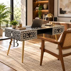 Könnte beinhalten: Ein rechteckiger Schreibtisch mit einem schwarz-weißen Blumenmuster-Inlay auf der Oberfläche und den Schubladenfronten. Der Schreibtisch steht auf vier schlanken, konisch zulaufenden goldfarbenen Metallbeinen und hat zwei Schubladen mit Knöpfen.