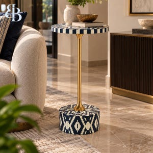 May include: A round side table featuring a navy blue and white geometric patterned top and base, connected by a slender gold-toned metal pedestal. The table surface holds a textured white vase and a small dark bowl resting on a book.