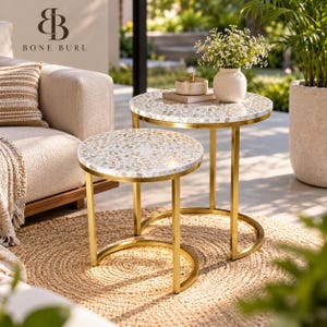 Könnte beinhalten: Zwei runde Satztische mit goldfarbenen Metallrahmen und weiß eingelegten Tischplatten, die ein Mosaikmuster aus kleinen, hellen Teilen aufweisen. Die Tische sind zusammen angeordnet, wobei der kleinere vor dem größeren steht.