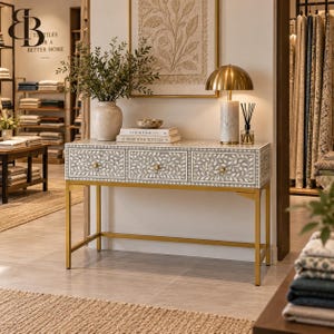 May include: A rectangular console table with three drawers featuring a grey and white floral inlay pattern, supported by a gold metal frame. The table holds a white vase with greenery, two stacked books, and a gold lamp with a marble base.