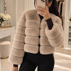 May include: A beige, cropped, long-sleeved jacket with a horizontal tiered, plush texture. The jacket features a collarless, open front design with a soft, dense appearance and fitted dark brown sleeves visible underneath the cuffs.
