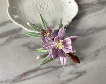 May include: A delicate hair comb with two purple flowers and green leaves. The flowers have yellow centers and are accented with small white pearls.